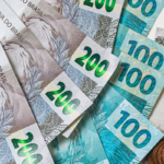 Imagem em close de cédulas e moedas de real brasileiro espalhadas sobre uma superfície clara. São visíveis notas de R$ 200, R$ 100, R$ 50 e R$ 20, além de moedas de diferentes valores empilhadas e agrupadas no lado direito da composição. A fotografia representa finanças, economia, recebimento de valores, investimentos ou planejamento financeiro, com foco nos detalhes e cores do dinheiro brasileiro.