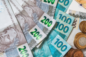 Imagem em close de cédulas e moedas de real brasileiro espalhadas sobre uma superfície clara. São visíveis notas de R$ 200, R$ 100, R$ 50 e R$ 20, além de moedas de diferentes valores empilhadas e agrupadas no lado direito da composição. A fotografia representa finanças, economia, recebimento de valores, investimentos ou planejamento financeiro, com foco nos detalhes e cores do dinheiro brasileiro.